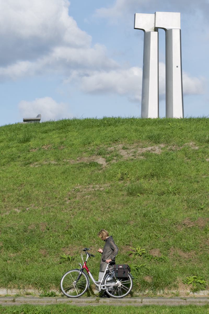 Woman standing by her bike checking her phone, in front of a hill with top of it the artwork Ver-kijkers by Bertie Kiewiet