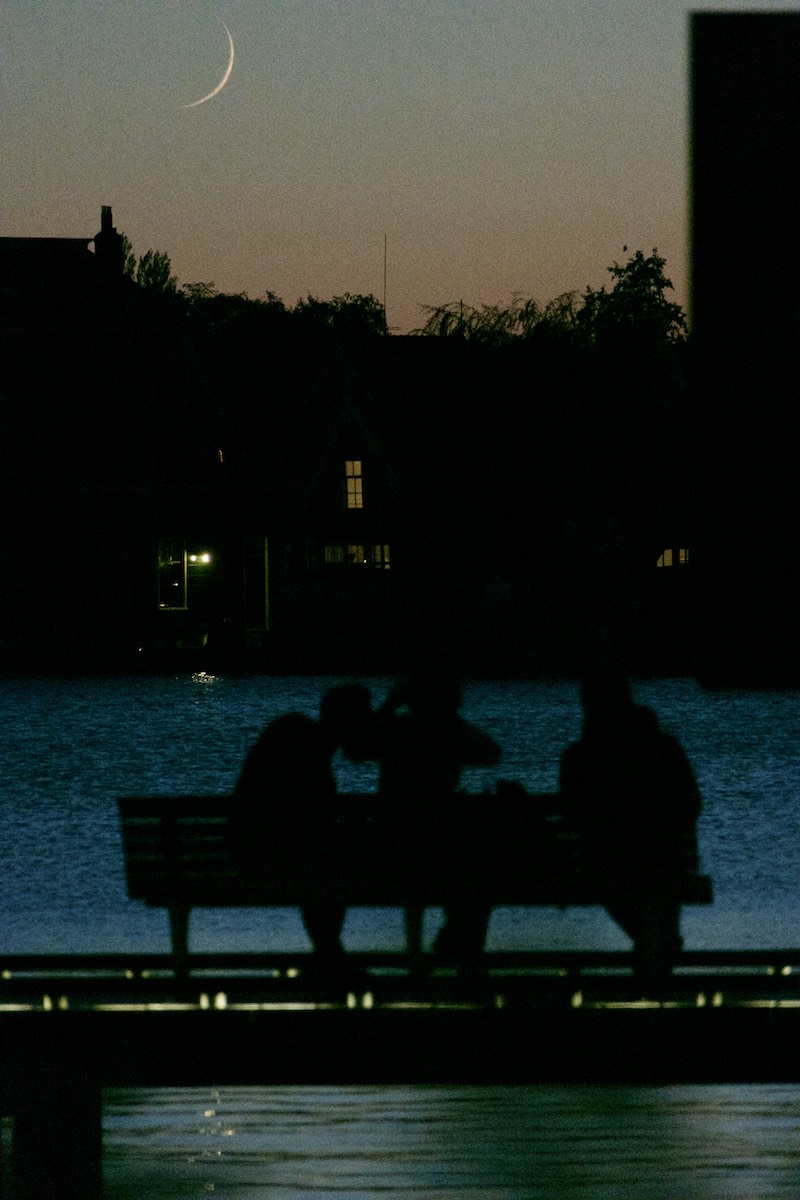 Zaan rover at near-dark. Setting crescent moon in the background across the river. Three people silhouetted on a bench in the foreground.