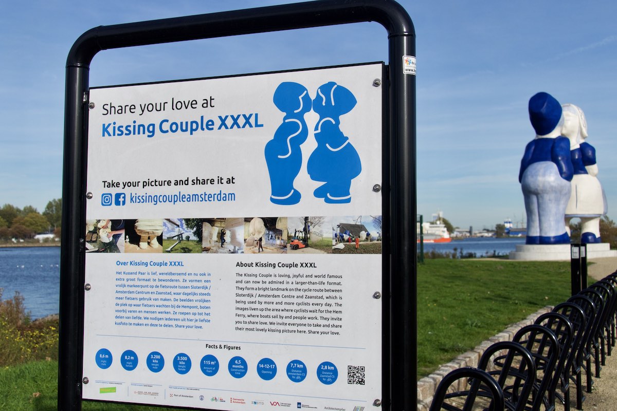 Sign describing the Kissing Couple XXXL artwork in the foreground, with the statue itself blurred in the background