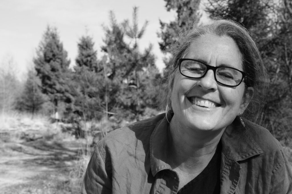 Black and white portrait photo of Abi. She is positioned on the right of the frame. She is smiling widely, showing teeth, with her eyes niched closed behind her rectangular-framed reading glasses.