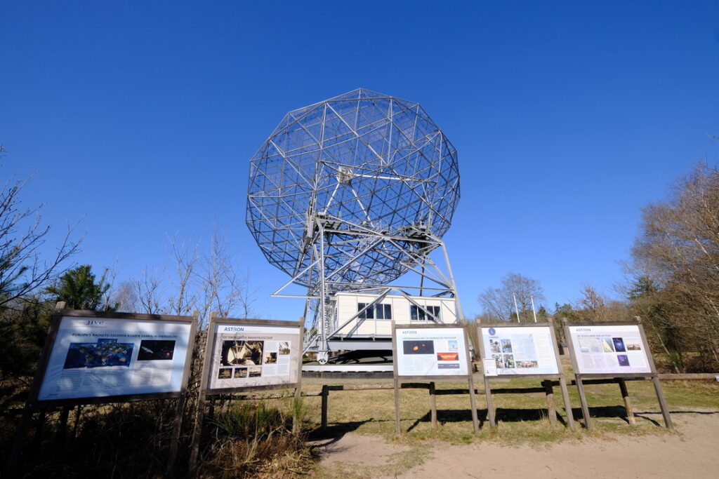 The radio telescope at Dwingelerveld against a clear blue sky. In the foreground are five display boards showing images from the construction, history, and scientific uses of the telescope. 