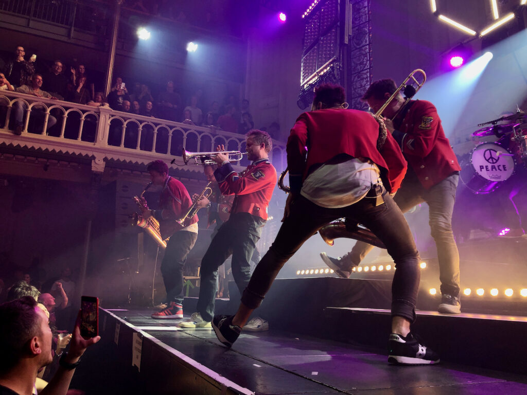 Meute on stage at Paradiso. The trumpet player is surrounded by sax and trombone players, in a dynamic pose.