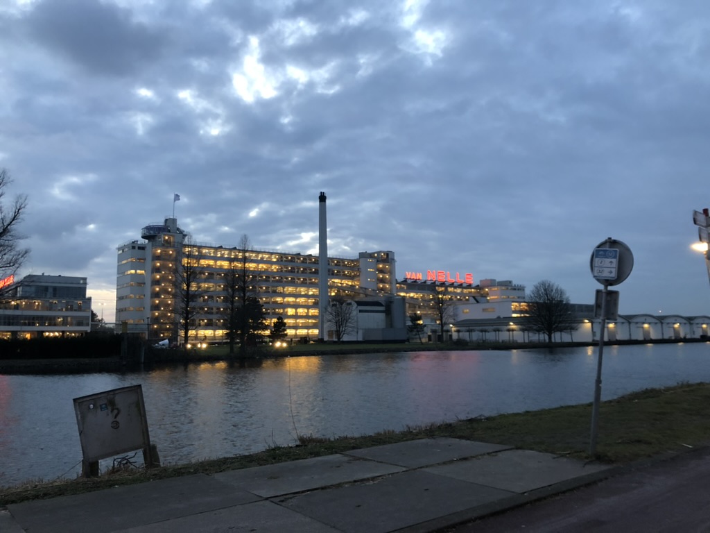 At dusk, a nine-storey factory building is lit up from the inside, on the other side of a wide canal. The words "Van Nelle" are lit up in red on the right of the building.