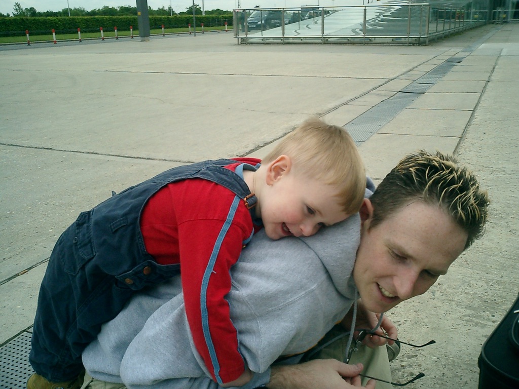 Martin kneeling down at the edge of a paved area, with Alex (two years old) on his back. Alex is wearing a red shirt and blue dungarees. They're both smiling. Martin's hair is dark, with bleach-blonde frosted tips.