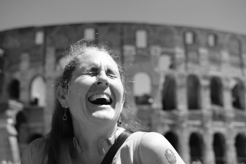 Abi with her eyes closed and her mouth open with laughter, lookup up towards the right of the frame . The Colosseum is behind her, out of focus, providing a soft blurry background.