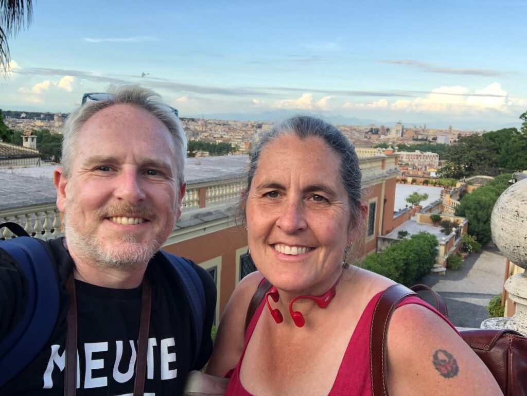 Martin and Abi looking at the camera from a balcony, with Rome stretching out in the distance behind them. Cumulus clouds draw a line above the horizon before the sky shades into a late-afternoon blue. Martin is wearing a black t-shirt with the white text MEUTE (as in the band Meute), and has a blue rucksack strapped on. Abi has her hair pulled back. She's wearing a red dress, and carring a brown leather backpack. Her red bone conduction headphones are around her neck.